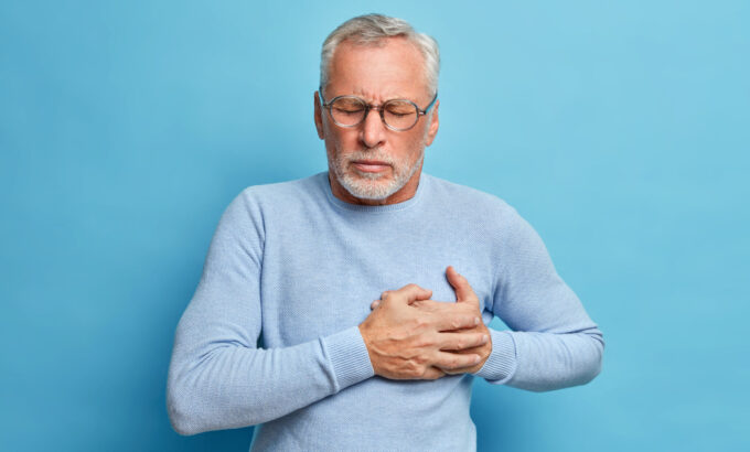 Senior man in spectacles presses hand to chest has heart attack suffers from unbearable pain closes eyes wears optical glasses poses against blue background. People age and problems with health